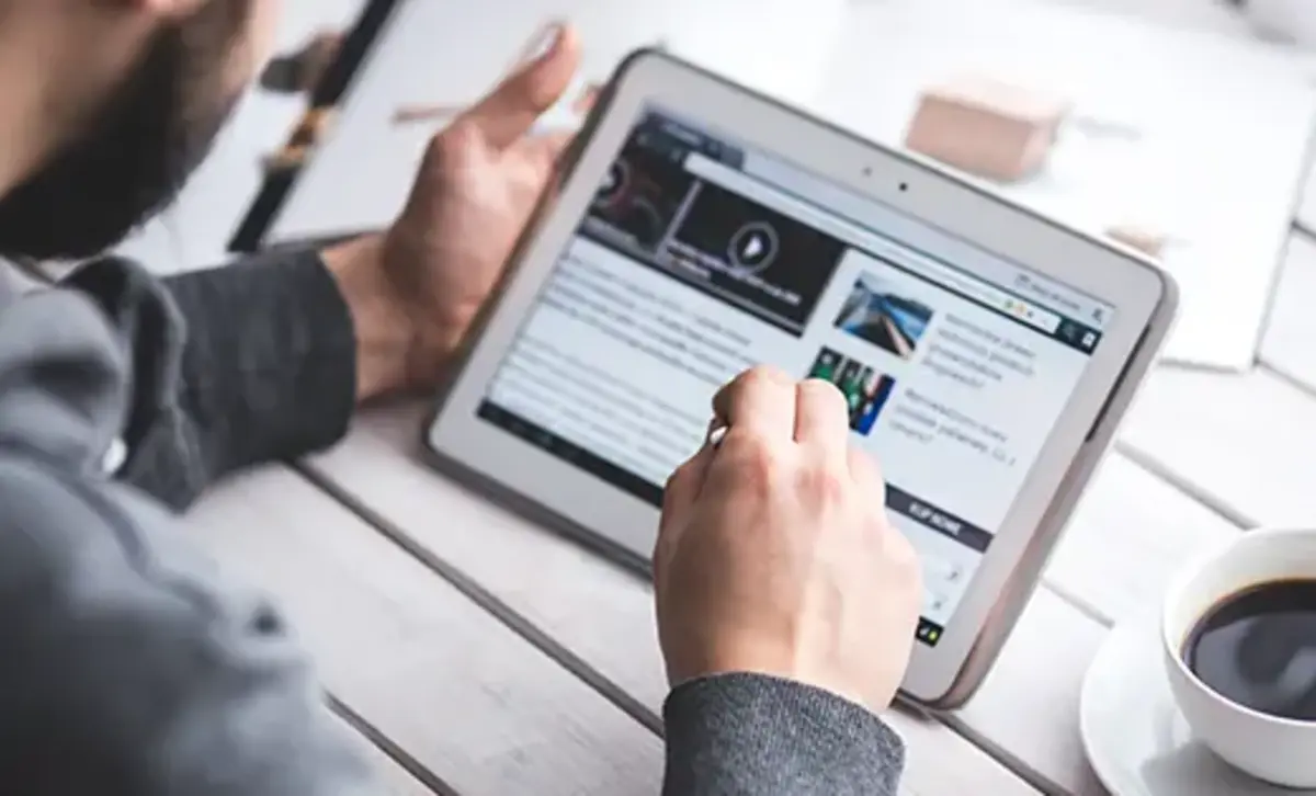A person using a tablet to browse a website or read content, with a cup of coffee nearby.