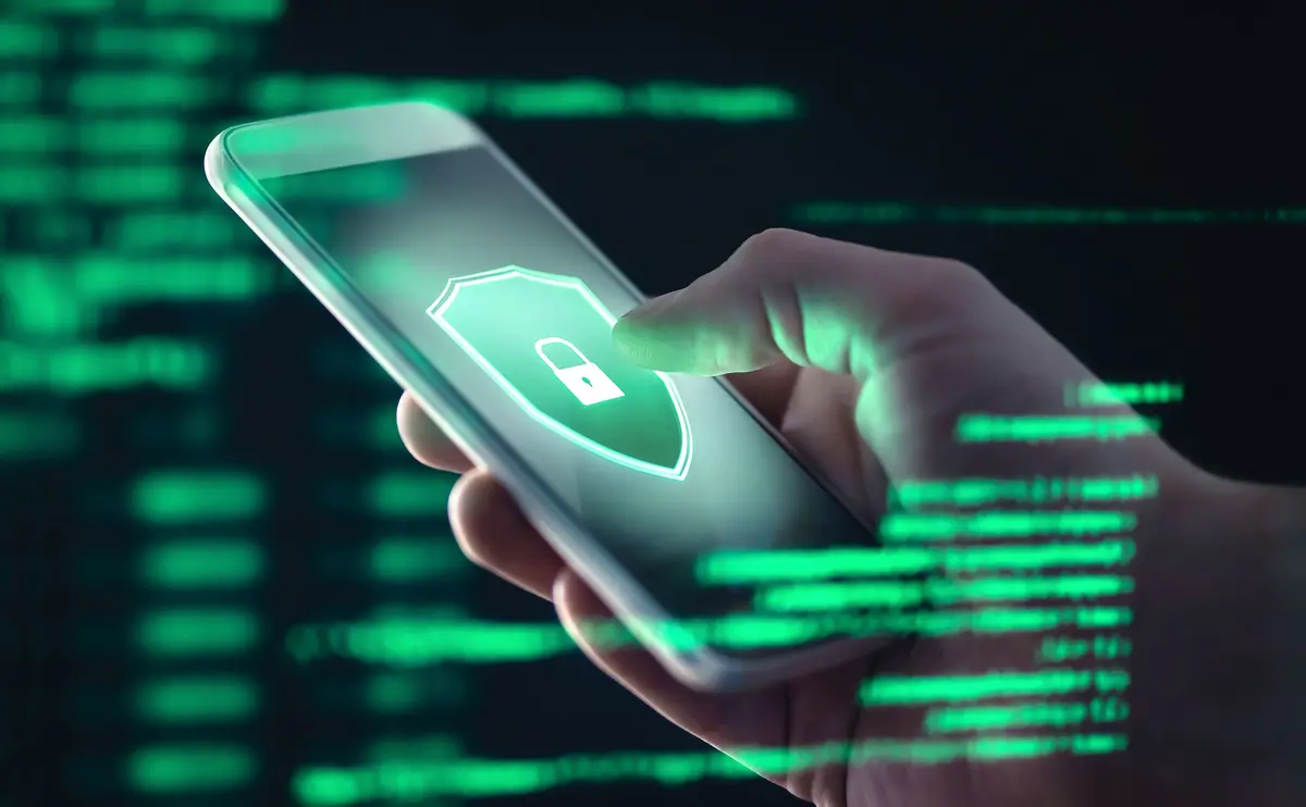 A hand holding a smartphone with a glowing shield icon featuring a padlock, symbolizing security or data protection.