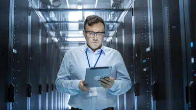 Les bases de la communication de crise. Un homme portant des lunettes, une chemise et une tablette marchant au milieu d'une salle de serveurs informatique.