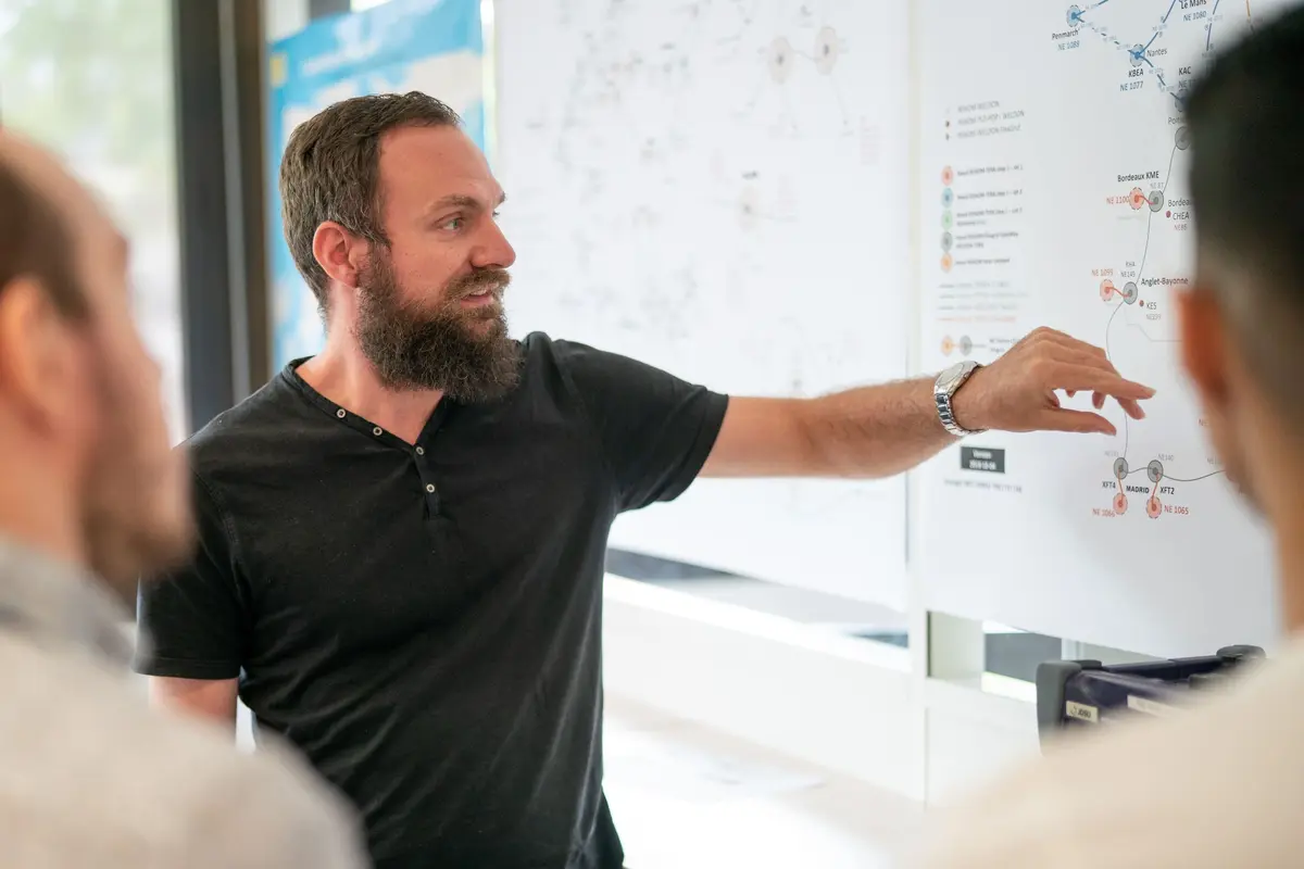 A man giving a presentation to a group, pointing at a large diagram on a whiteboard, in a professional setting.