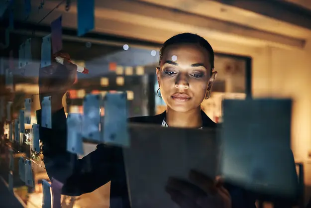 Professional working late, planning on a glass wall with sticky notes while holding a tablet