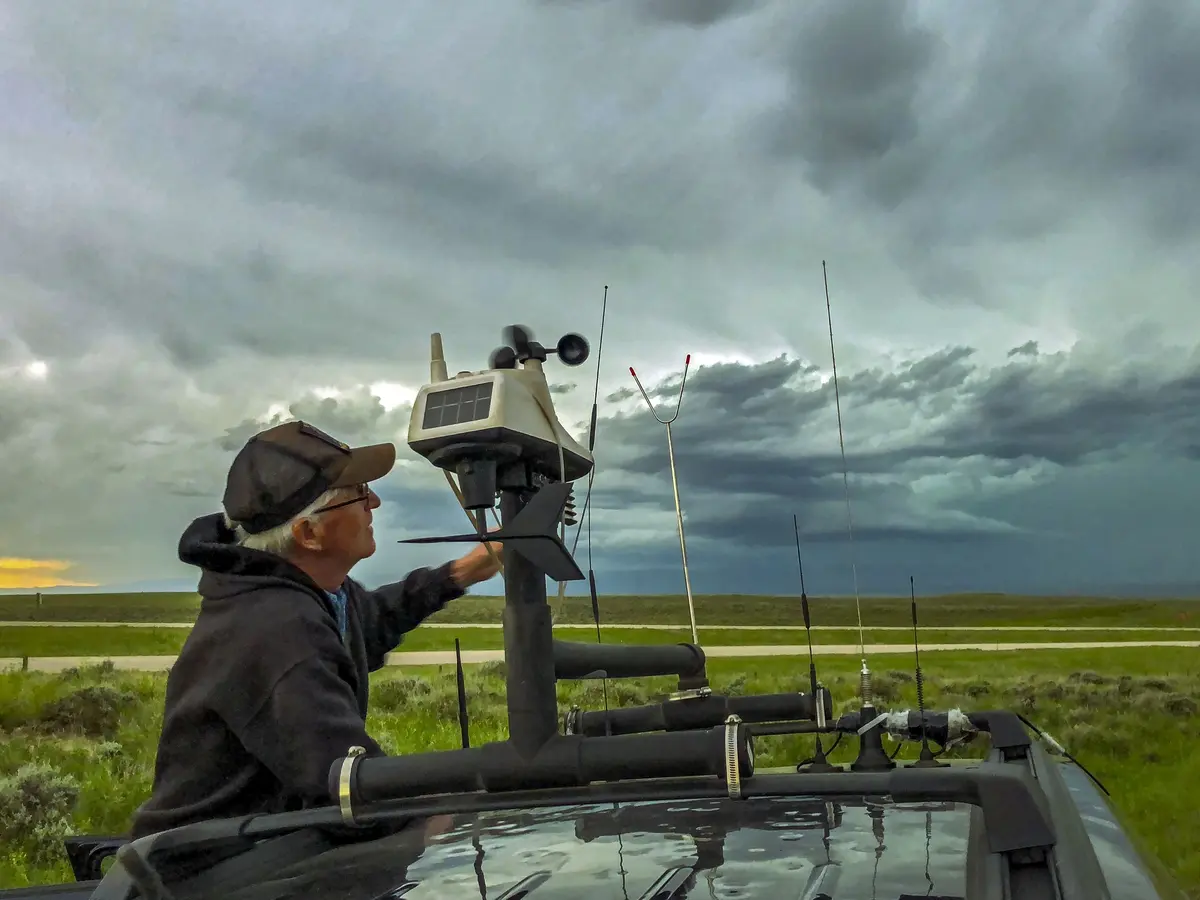 A person operating a weather or atmospheric monitoring station mounted on a vehicle, with a dramatic, stormy sky in the background.