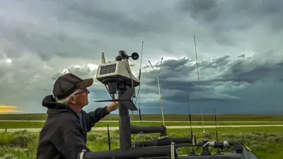 A person operating a weather or atmospheric monitoring station mounted on a vehicle, with a dramatic, stormy sky in the background.