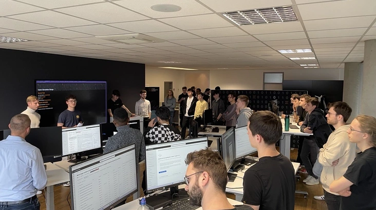 Gruppe von Studierenden steht in einem modernen Büro mit mehreren Computerarbeitsplätzen und großen Monitoren, während zwei Personen vorne eine Präsentation halten und die Zuhörenden aufmerksam zuhören.