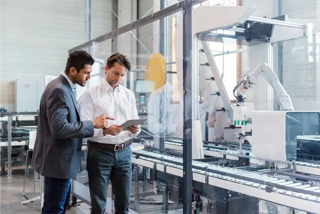 Two professionals reviewing a tablet next to an automated production line with a robotic arm