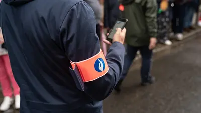 Person wearing a dark jacket with a red armband, standing among a crowd, possibly at an event or protest.