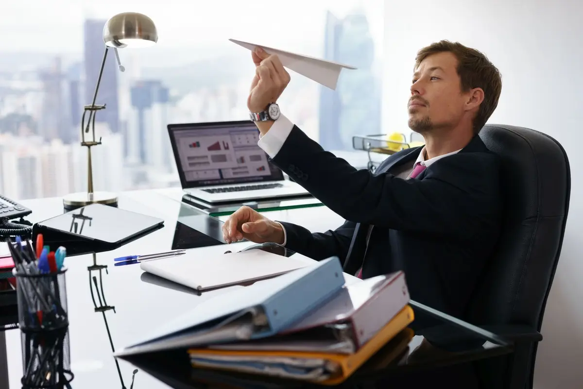 A man sitting at a cluttered office desk, holding a paper airplane and looking thoughtfully into the distance.