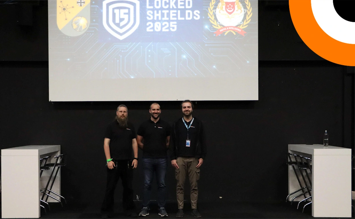 Orange Cyberdefense présent à Locked Shields 2025 Trois professionnels de la société Orange Cyberdefense se tenant côte à côte pendant un événement organisé par l'OTAN.