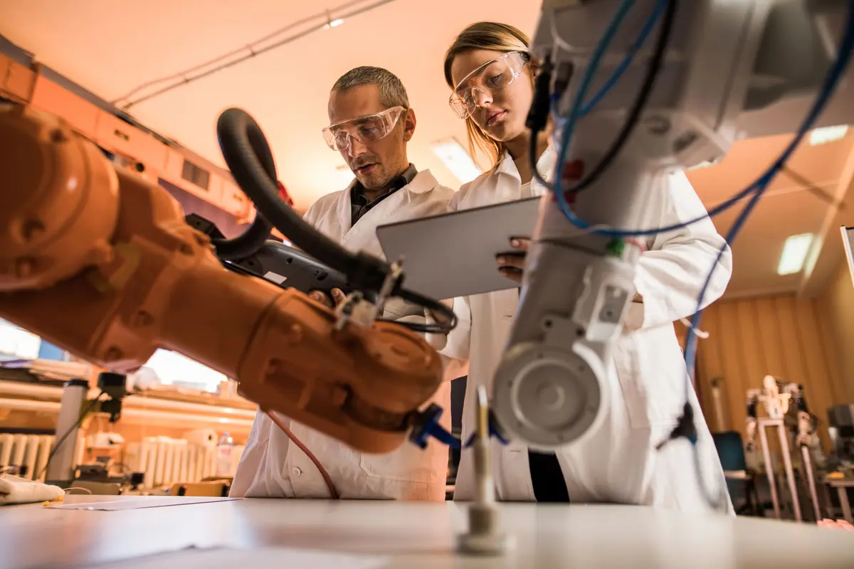 Two scientists or engineers working together in a laboratory or industrial setting, examining a robotic arm or automated machinery.
