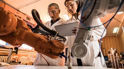Two scientists or engineers working together in a laboratory or industrial setting, examining a robotic arm or automated machinery.