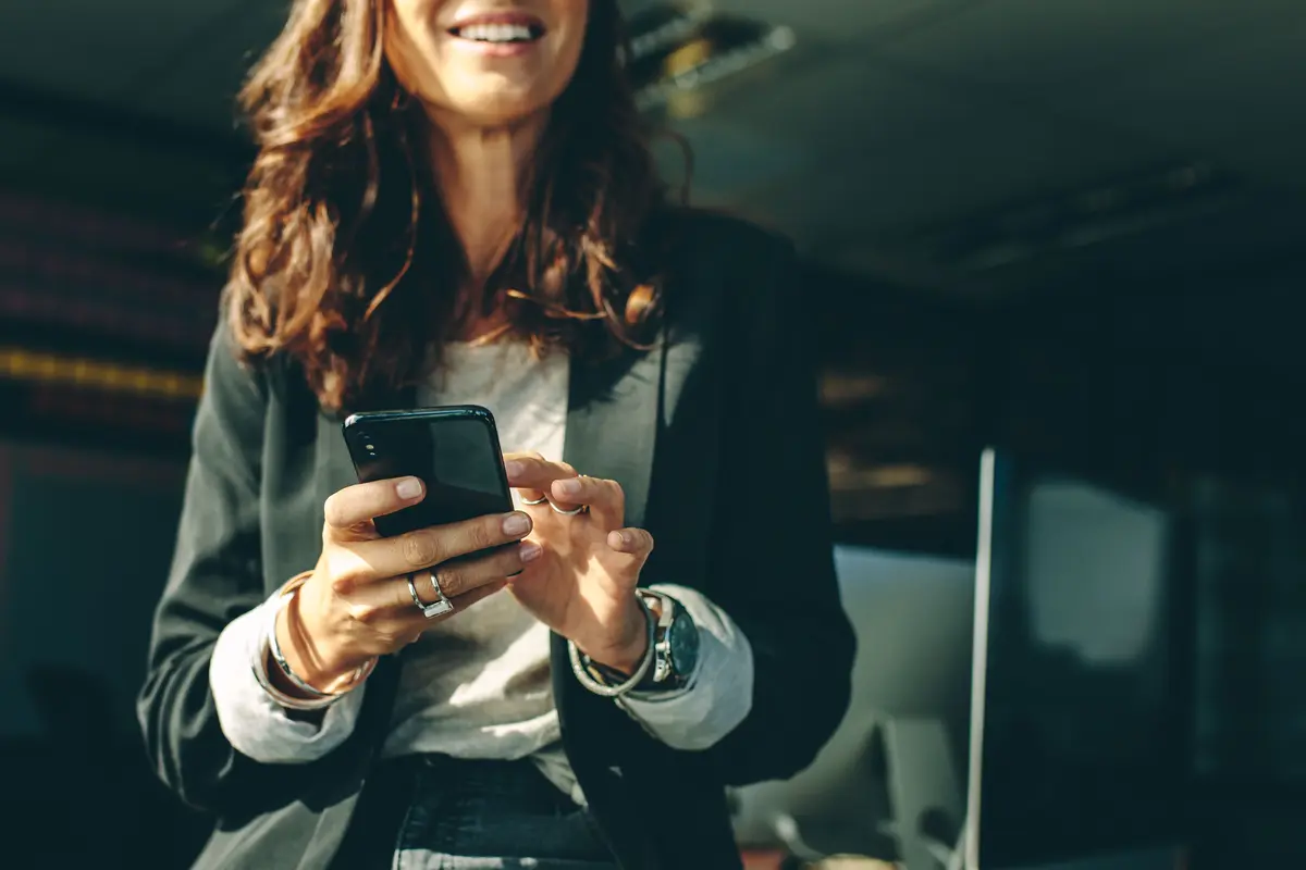 A woman smiling while using her smartphone in an office or professional environment.