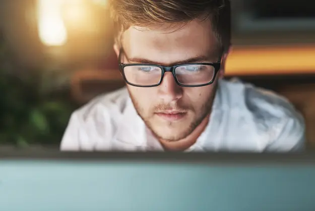 IT professional with glasses focused on laptop screen in warm office lighting